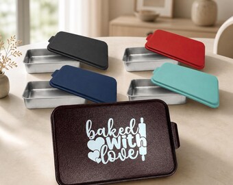 May include: A set of rectangular metal baking pans with lids in black, red, navy blue, and teal. One dark brown lid features the white text "baked with love" alongside a heart graphic and a rolling pin icon, positioned in the foreground.