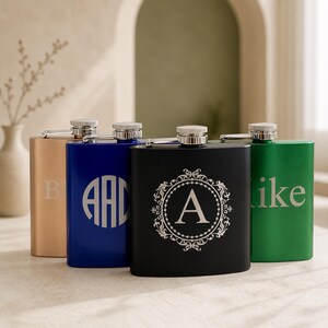 May include: Four rectangular metal flasks with screw-top caps in gold, blue, black, and green. The gold flask features the letter B, the blue flask has a circular monogram AAD, the black flask displays a letter A in a filigree frame, and the green flask shows the text like.