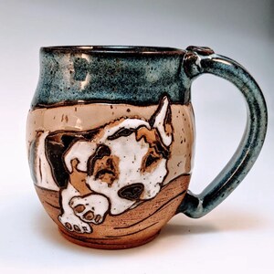 May include: A ceramic mug with a brown, white, and black dog design. The mug has a blue glaze and a brown handle.