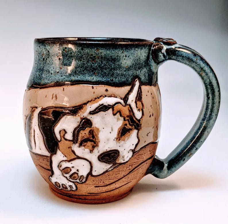 May include: A ceramic mug with a brown, white, and black dog design. The mug has a blue glaze and a brown handle.