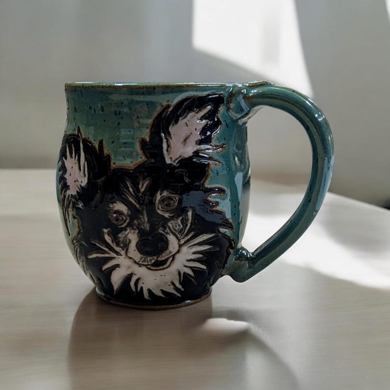 May include: A teal ceramic mug with a black and white dog face illustration. The dog has white fur around its face and ears.