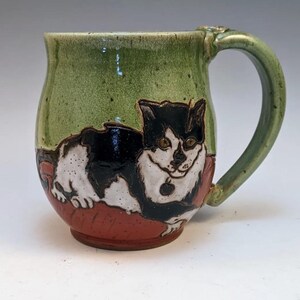 May include: A green ceramic mug with a black and white cat illustration. The cat is lying on a red pillow. The mug has a handle.