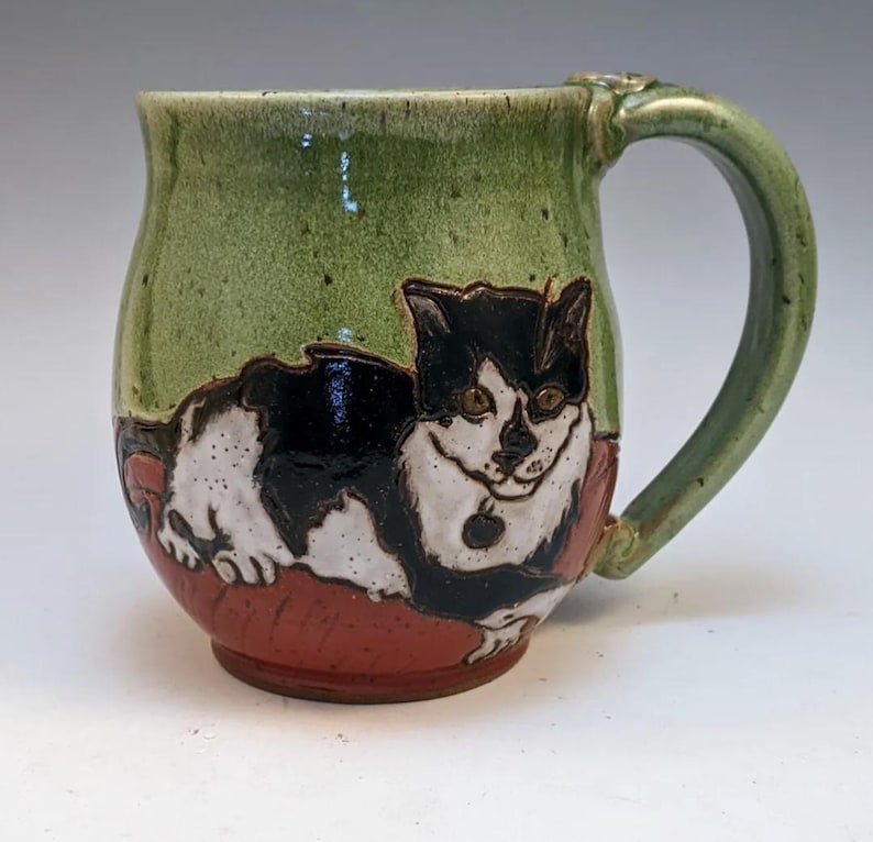 May include: A green ceramic mug with a black and white cat illustration. The cat is lying on a red pillow. The mug has a handle.