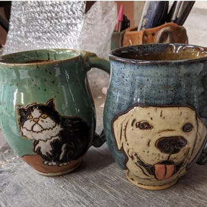 Puede incluir: Dos tazas de cerámica con ilustraciones de animales. La taza de la izquierda es verde con una ilustración de un gato blanco y negro. La taza de la derecha es azul con una ilustración de un perro blanco.