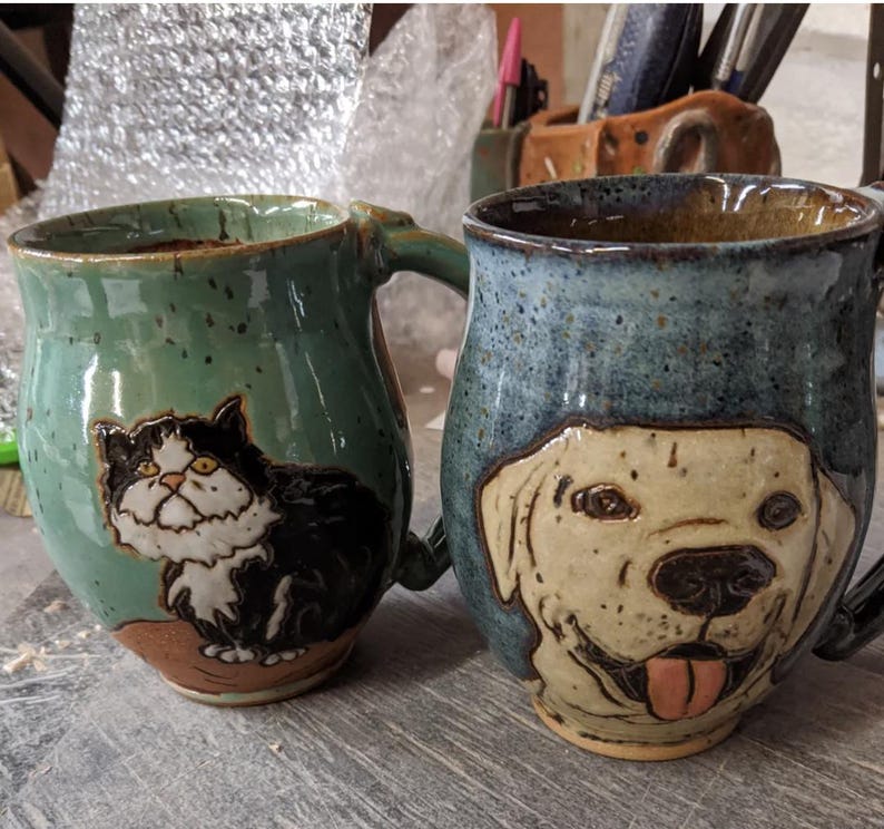 May include: Two ceramic mugs with animal illustrations. The mug on the left is green with a black and white cat illustration. The mug on the right is blue with a white dog illustration.