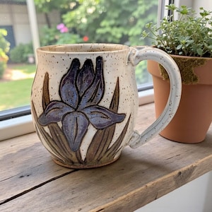 May include: A handmade ceramic mug with a speckled, off-white glaze. The mug features a large, stylized iris flower in shades of blue and brown. The handle is curved and the mug sits on a wooden surface.
