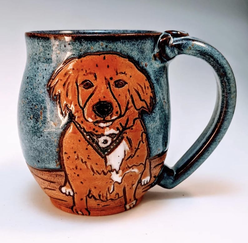 May include: A blue and brown ceramic mug with a hand-painted image of a dog. The dog is brown and white with a bandana around its neck. The mug has a rounded body and a handle.