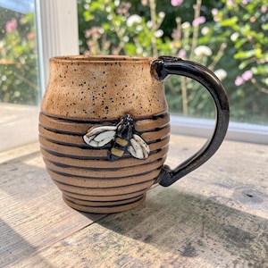 May include: A ceramic mug with a speckled tan upper section and a ribbed lower body featuring horizontal dark brown lines. A sculpted bee with white wings and yellow and black stripes is attached to the side. It has a dark brown handle.