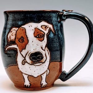May include: A ceramic mug with a hand-painted image of a dog. The dog is white with brown markings and has a black nose. The mug is blue and brown with a speckled glaze.