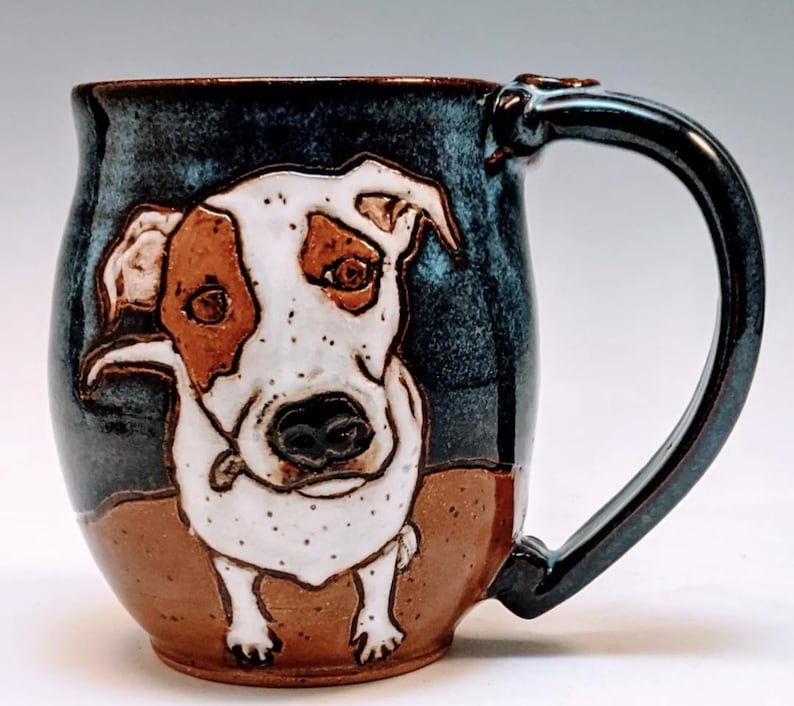 May include: A ceramic mug with a hand-painted image of a dog. The dog is white with brown markings and has a black nose. The mug is blue and brown with a speckled glaze.