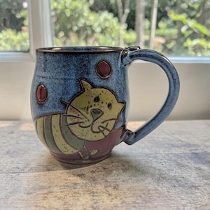 Puede incluir: Una taza de cerámica hecha a mano con un esmalte azul y un diseño caprichoso de gato. El gato es amarillo y verde con un collar rojo. La taza tiene una forma redondeada y un asa grande. La taza mide aproximadamente 10 cm de alto.