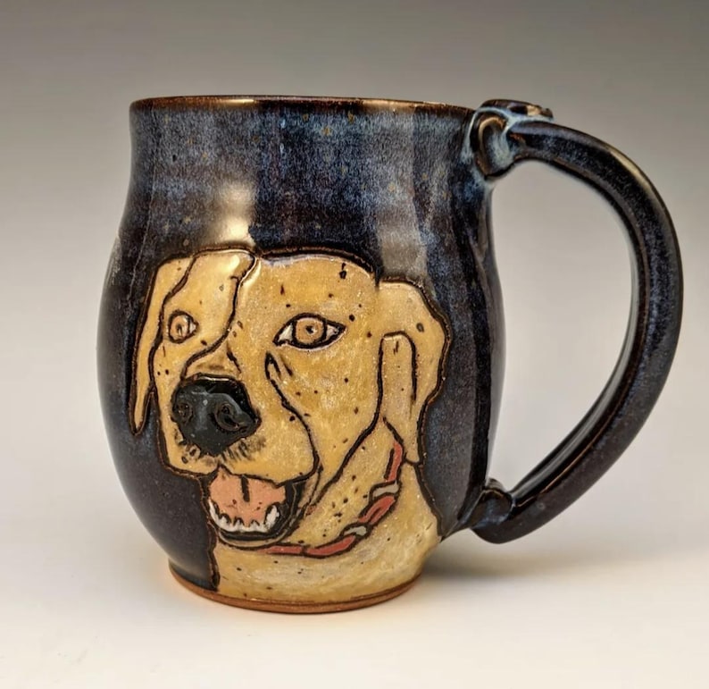 May include: A ceramic mug with a hand-painted image of a yellow Labrador Retriever's face. The mug has a dark blue glaze with brown accents and a brown handle.