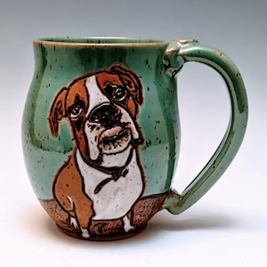 May include: A green ceramic mug with a brown and white boxer dog illustration. The mug has a speckled glaze and a rounded body.