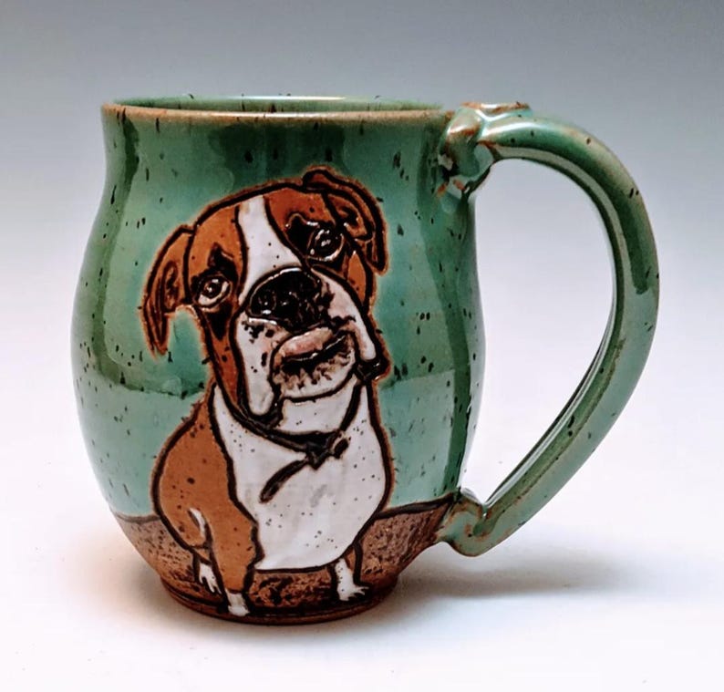 May include: A green ceramic mug with a brown and white boxer dog illustration. The mug has a speckled glaze and a rounded body.