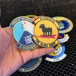 May include: A collection of five round, gold-colored patches with various designs and text. The patches feature a blue and white patch with the text "Force of the North" and "3D Maintenance", a yellow patch with the text "8th Fighter SQ" and "The Black Sheep", a blue and white patch with the text "65th Communications", a yellow patch with the text "8 SOS", and a blue and white patch with the text "US Air Force" and "United States of America".