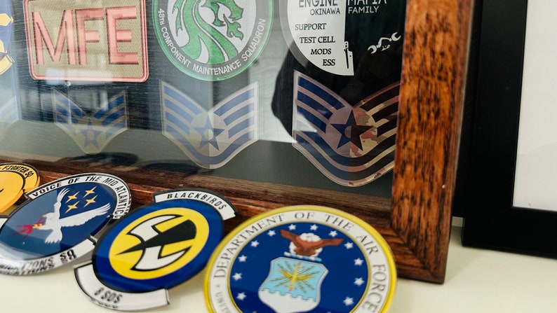 May include: A framed display of military patches and insignia, including a green patch with a dragon, a blue patch with a star, and a blue and yellow patch with a lightning bolt. The frame is made of dark wood and has a glass front.