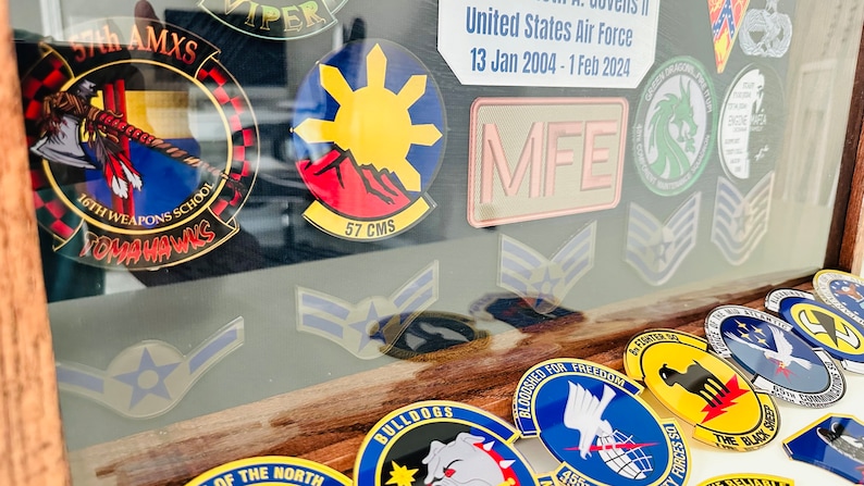 May include: A framed display of military patches and insignia, including a yellow and red patch with a sun and mountains, a green patch with a dragon, and a gray patch with the letters "MFE".