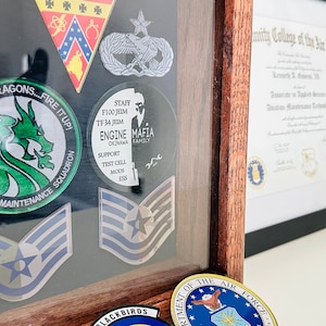 May include: A wooden shadow box with a variety of military patches and a diploma from the Community College of the Air Force. The patches include a green dragon, a blue and white star, and a black and white patch with the words "Mafia Family". The diploma is framed in black and has a gold seal.
