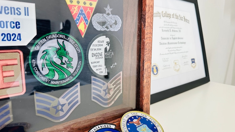 May include: A wooden shadow box with a variety of military patches and a diploma from the Community College of the Air Force. The patches include a green dragon, a blue and white star, and a black and white patch with the words "Mafia Family". The diploma is framed in black and has a gold seal.