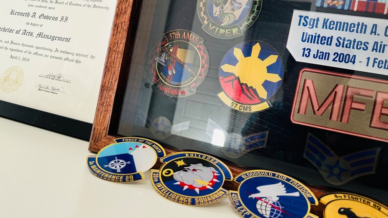 May include: A framed display of military patches and pins. The patches are mostly blue, red, and yellow with gold trim. The pins are mostly gold with blue and red accents. The display includes the text "57th AMXS", "VIPER", "57 CMS", "FORCE OF THE NORTH", "BULLDOGS", "35th INTELLIGENCE SQUADRON", "BLOODHOUND FOR FREEDOM", "455th EXP", and "8th FIGHTER SQ".