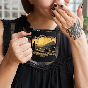 May include: Black ceramic mug featuring a golden landscape design with a wizard silhouette, full moon, and birds. The mug has a black handle and a decorative golden border. The design evokes a fantasy theme.