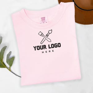 May include: A pink t-shirt with a black logo that says "YOUR LOGO HERE" and features two crossed paintbrushes.