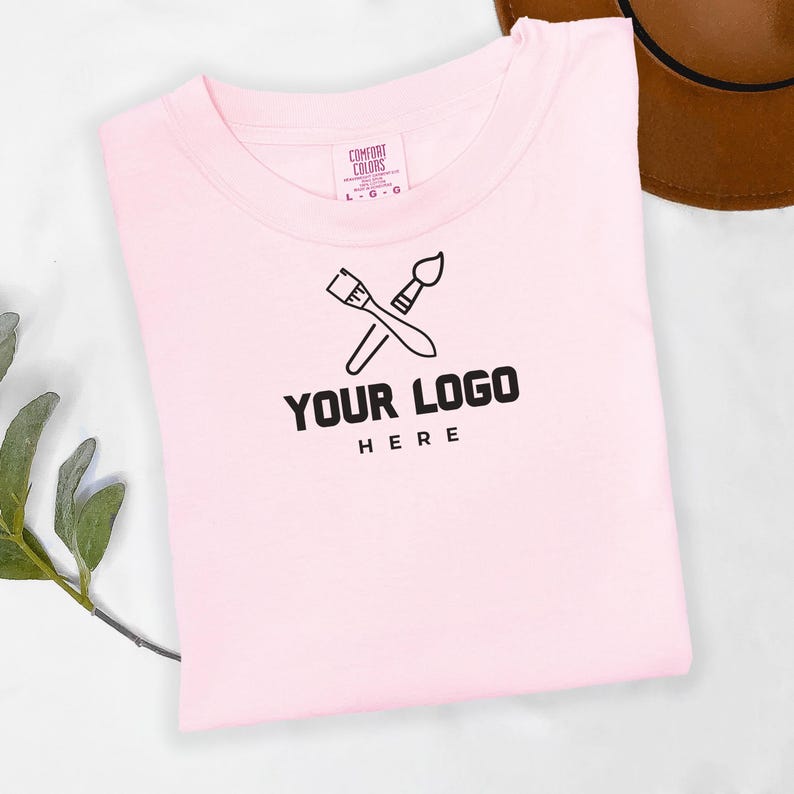 May include: A pink t-shirt with a black logo that says "YOUR LOGO HERE" and features two crossed paintbrushes.