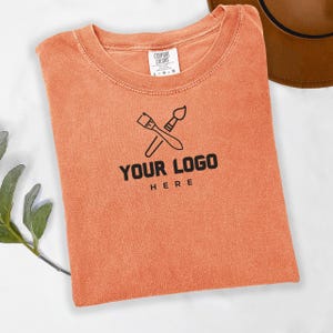 May include: A burnt orange t-shirt with a paintbrush and palette knife design and the text "YOUR LOGO HERE".