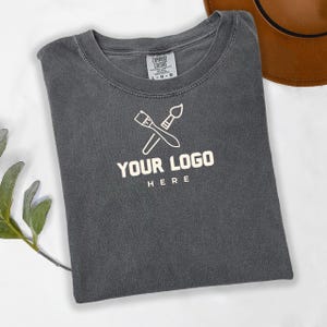 May include: A dark gray t-shirt with a white paintbrush and palette knife logo. The logo says "YOUR LOGO HERE".