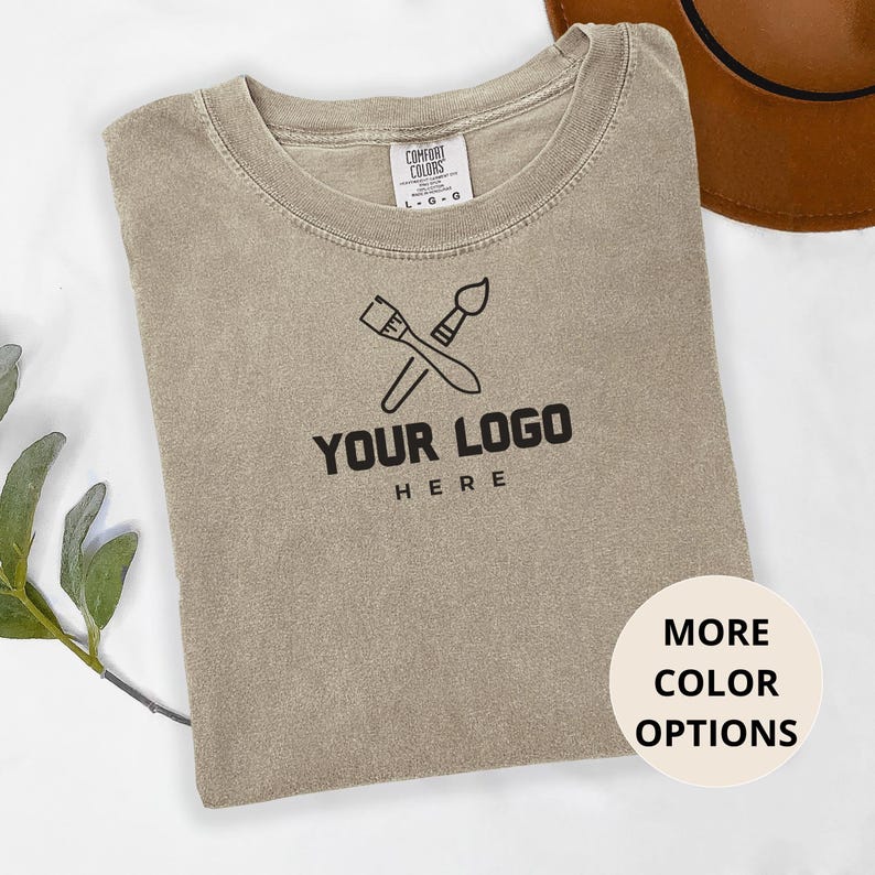 May include: A light brown t-shirt with a paintbrush and palette design and the text "YOUR LOGO HERE" printed on the front. The shirt is made of a soft, comfortable material and is available in a variety of colors.