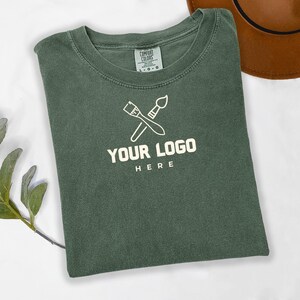 May include: A green t-shirt with a white paintbrush and paint roller design. The text "YOUR LOGO HERE" is printed below the design.