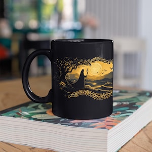 May include: Black ceramic mug featuring a gold illustration of a robed figure under a tree, with a full moon and birds in the background. The design includes intricate gold detailing and a decorative border. The mug is sitting on a book.