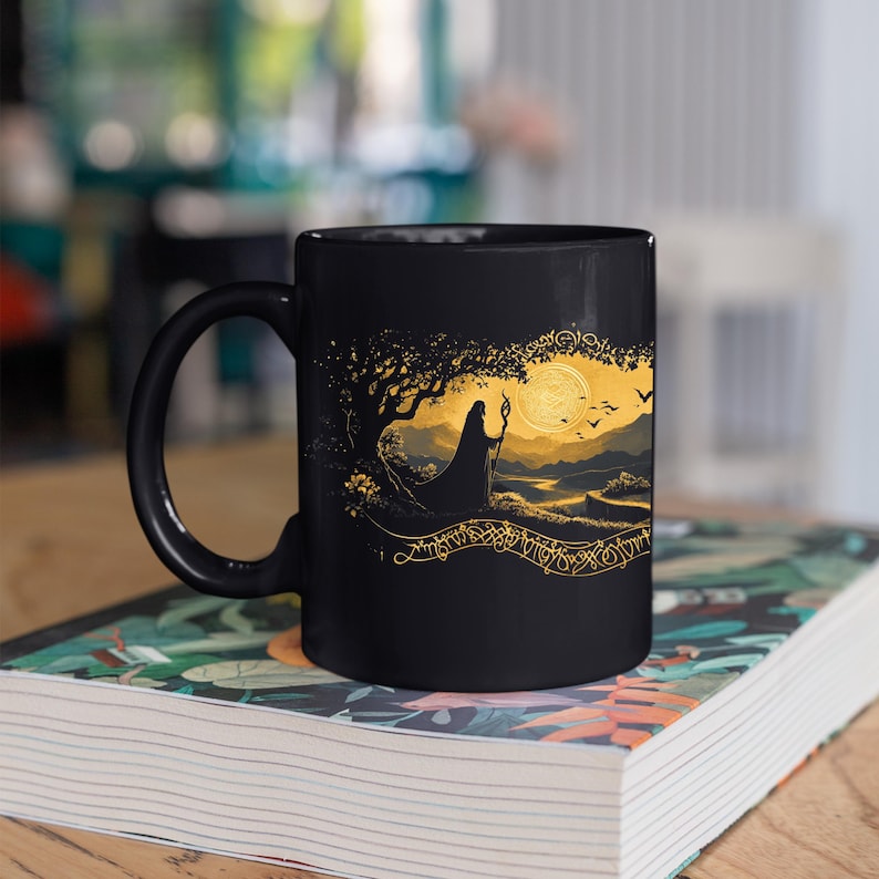May include: Black ceramic mug featuring a gold illustration of a robed figure under a tree, with a full moon and birds in the background. The design includes intricate gold detailing and a decorative border. The mug is sitting on a book.
