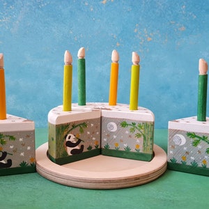 Op de afbeelding: Een houten speelgoedtaart met groene en witte frosting, versierd met bamboe en een panda. De taart heeft zes gele en groene kaarsen.