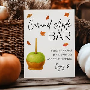 May include: A white sign with the words "Caramel Apple BAR" and instructions for creating caramel apples. The sign is decorated with fall leaves and an illustration of a caramel apple. Pumpkins and a basket are in the background.