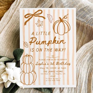 Puede incluir: Una tarjeta de invitación con temática otoñal con el texto "A Little Pumpkin Is On The Way!". La tarjeta presenta ilustraciones de calabazas y está decorada con un lazo, hojas y un diseño de calabaza apilada. La tarjeta está sobre una tela beige.