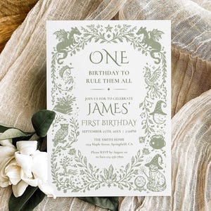 May include: A white invitation card with green illustrations and text, including the words "ONE BIRTHDAY TO RULE THEM ALL" and "JAMES' FIRST BIRTHDAY". The card is decorated with floral and fantasy-themed illustrations. The card is placed on a beige fabric with white flowers.