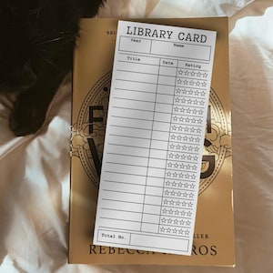 May include: A white library card with the text "LIBRARY CARD" is placed on top of a gold book. The card has sections for title, date, and rating, with star ratings. A black cat is partially visible in the upper left corner.
