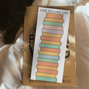 May include: A bookmark with the text "2026 READING LIST" and a stack of colorful book illustrations rests on a gold-colored book. The bookmark features pastel colors, including pink, green, and yellow.