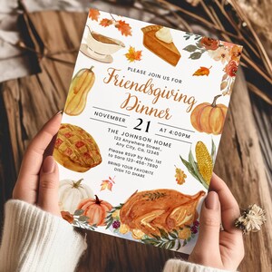 Op de afbeelding: Een Friendsgiving Dinner-uitnodiging die door een persoon wordt vastgehouden. De uitnodiging bevat aquarel illustraties van Thanksgiving-eten, waaronder een gebraden kalkoen, pompoenen en taart. De tekst luidt "Friendsgiving Dinner" en bevat de datum, tijd en locatie.