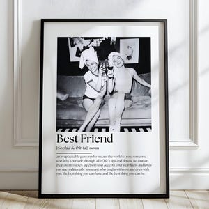 May include: Black and white framed print featuring a vintage photograph of two women toasting with glasses. The text "Best Friend" is displayed below the image, along with a definition. The frame is black, and the print is displayed against a white wall.