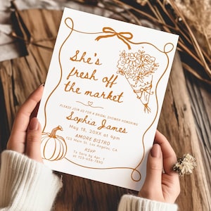 May include: A bridal shower invitation with the text "She's fresh off the market". The invitation features details for Sophia James' shower, with a pumpkin and floral bouquet illustration in a warm, earthy colour scheme. The invitation is held by a person.