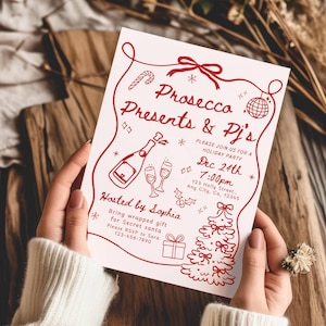 Op de afbeelding: Een uitnodiging voor een kerstfeest met rode illustraties en tekst. De uitnodiging luidt "Prosecco, Presents & Pj's" en geeft de datum 24 december om 19.00 uur. De uitnodiging wordt vastgehouden door een persoon die een witte trui draagt.