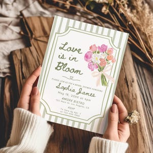 May include: A bridal shower invitation with a floral bouquet illustration. The invitation features the text "Love is in Bloom" and details for Sophia James' shower, including the date, time, and location. The design has a striped border and a soft colour palette.