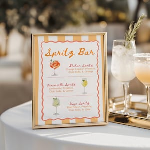 May include: A gold-framed sign with the words "Spritz Bar" in orange script. The sign lists three spritz cocktail recipes: Italian, Limoncello, and Hugo, each with watercolor illustrations of the drinks. The sign is on a white table.