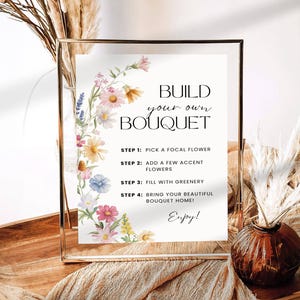 May include: A gold-framed sign with the text "Build Your Own Bouquet" and instructions, decorated with watercolour flowers. The sign is on a wooden tray with dried plants and a vase with dried flowers.