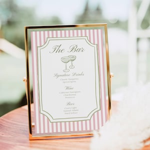 May include: A gold framed sign with a pink and white striped background lists "The Bar" and "Signature Drinks" including cocktails, wine, beer, and hard seltzer. The sign is displayed on a wooden tray.