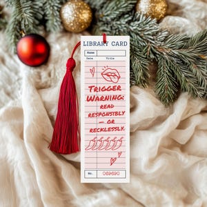 May include: A rectangular bookmark with a red tassel, featuring a library card design. The bookmark has the words "TRIGGER WARNING: READ RESPONSIBLY - OR RECKLESSLY." printed on it, along with a number and some small illustrations. The background includes Christmas decorations.