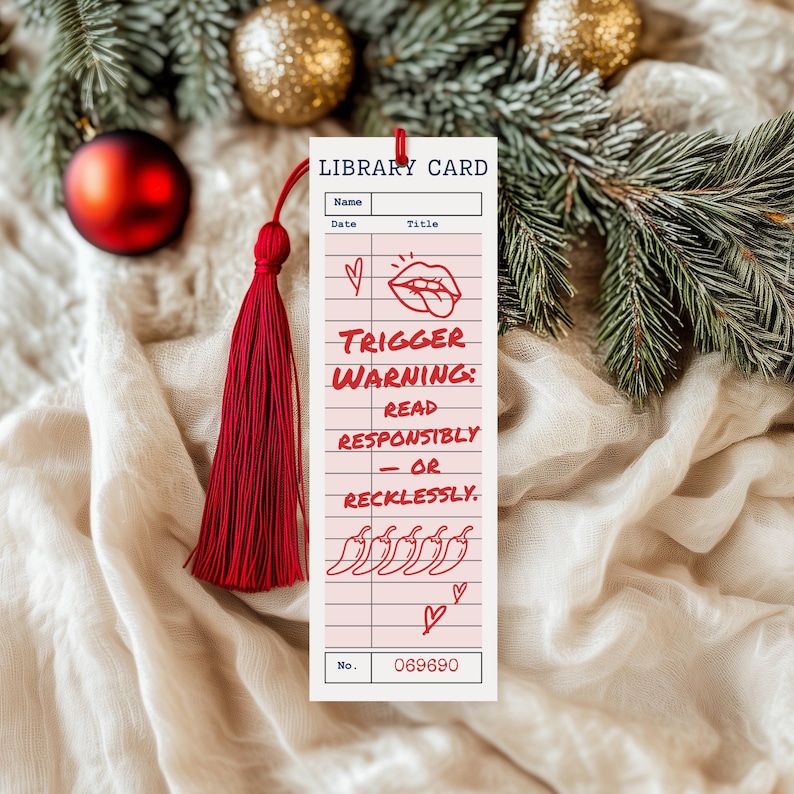 May include: A rectangular bookmark with a red tassel, featuring a library card design. The bookmark has the words "TRIGGER WARNING: READ RESPONSIBLY - OR RECKLESSLY." printed on it, along with a number and some small illustrations. The background includes Christmas decorations.