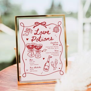 May include: A framed sign with the words "Love Potions" in red script. The sign lists signature drinks, wine, beer, and non-alcoholic options. The frame is gold, and the sign is on a wooden surface.